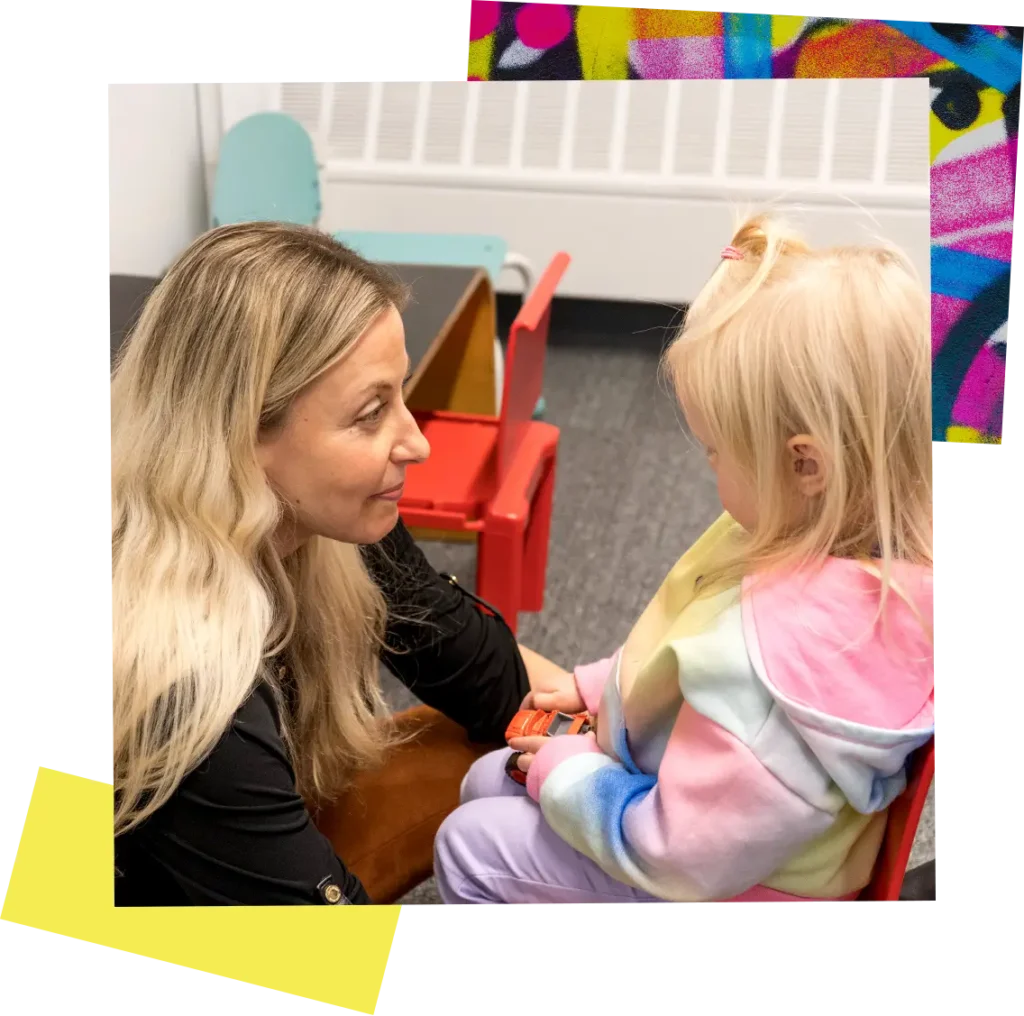 A small child plays with a toy car in a classroom setting while a woman studies her expression.